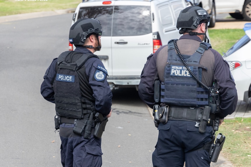 Two police officers in tactical gear stand on a street with their backs to the camera.