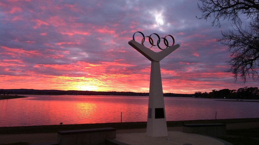 A monument featuring the Olympic rings in front of a lake, with a pink and yellow sunset in the background.