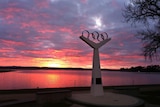 A monument featuring the Olympic rings in front of a lake, with a pink and yellow sunset in the background.