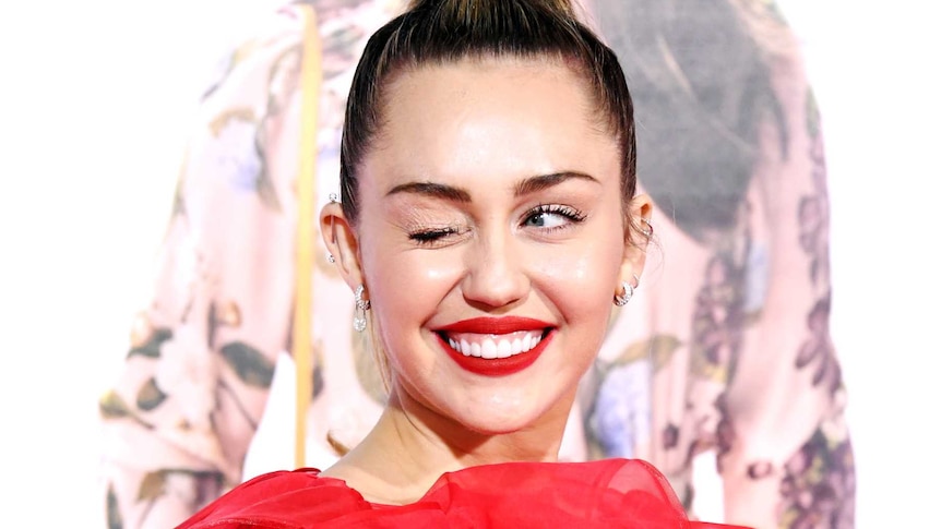 Miley Cyrus, wearing a red dress and lipstick, winks as she smiles for a photo.