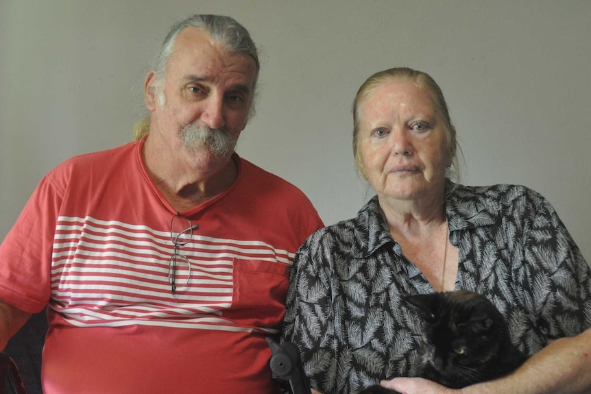 An elderly couple sit on the couch looking upset.