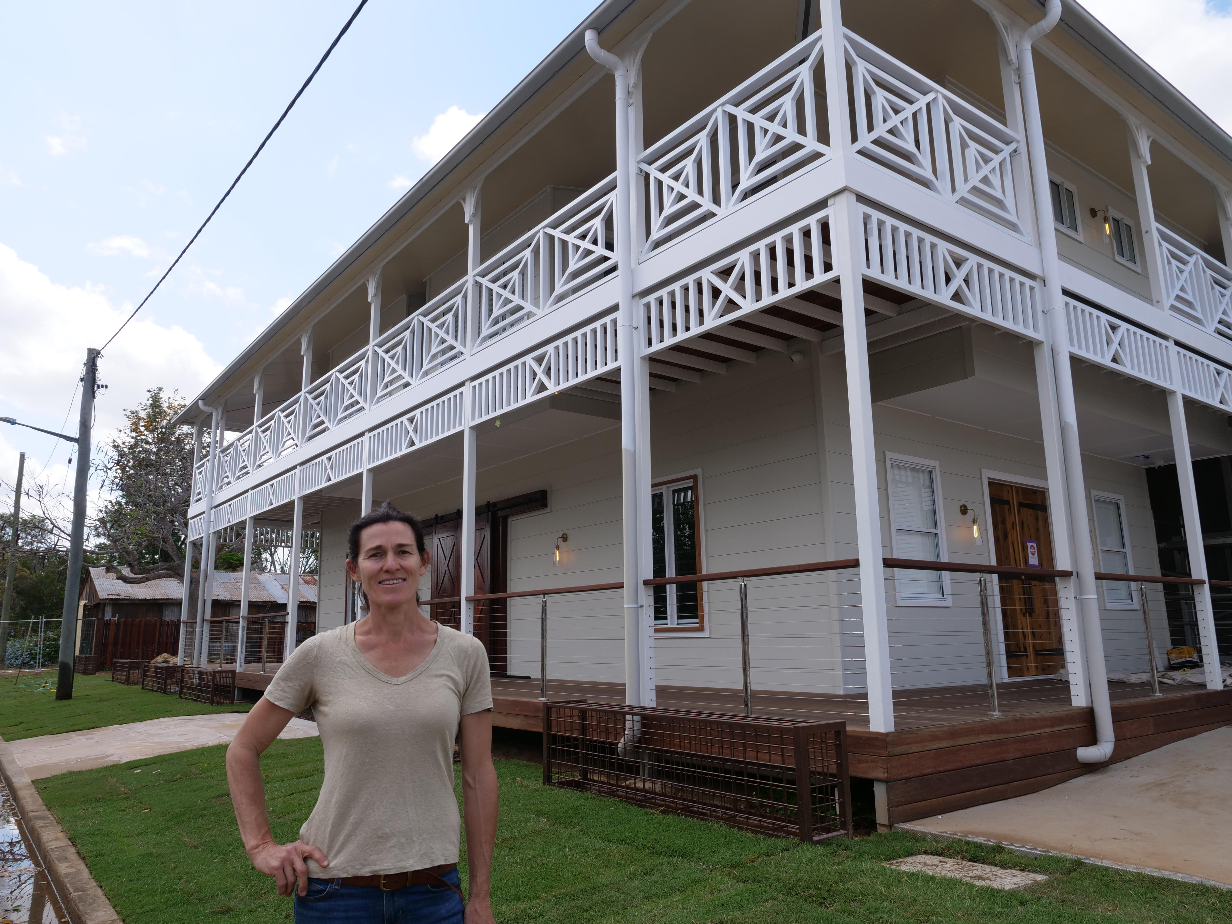 A woman with a brown ponytail, jeans and a tshirt stands in front of a freshly painted white and cream Queenslander-style pub