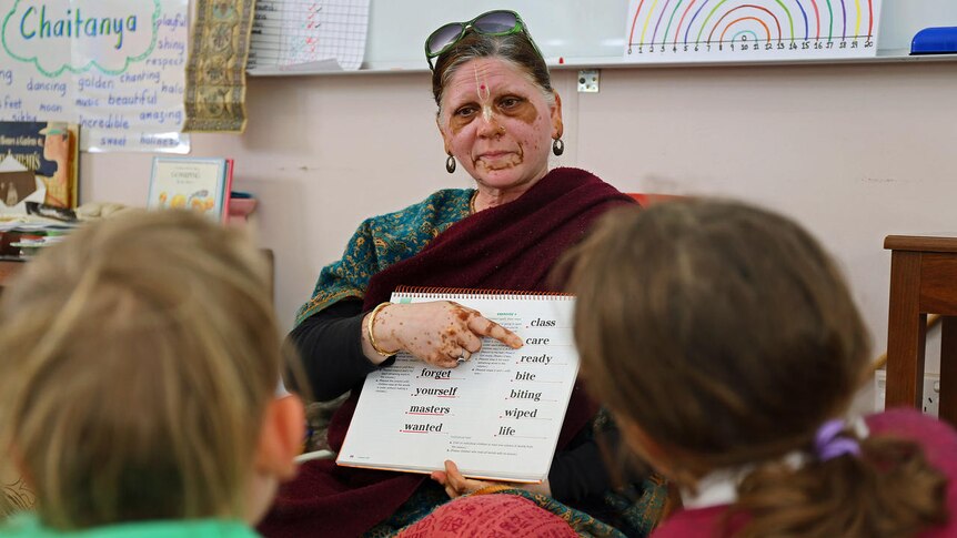 A woman in a sari and facepaint holds a book up in front of a group of students.
