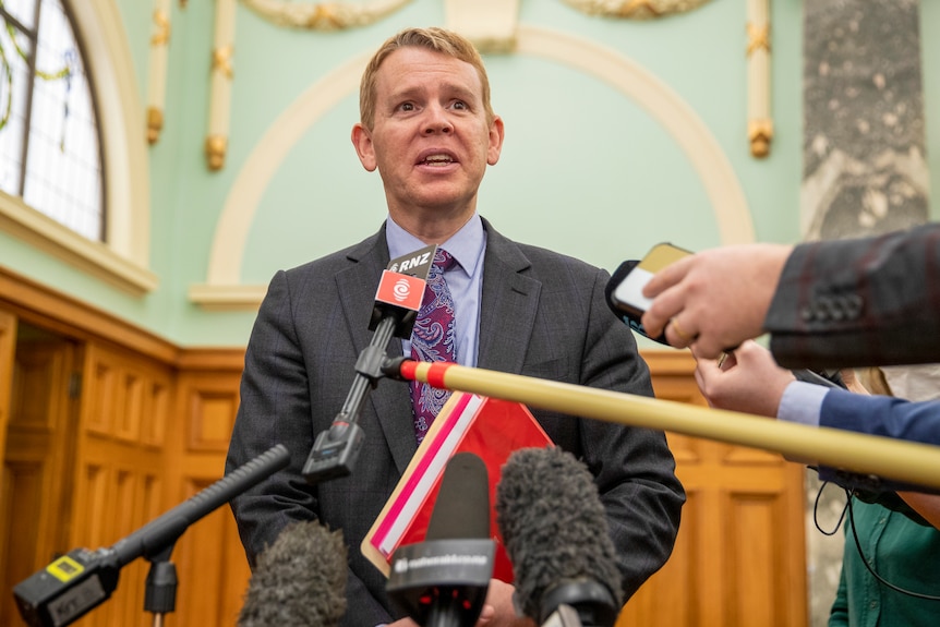 New Zealand Police Minister Chris Hipkins, wearing a suit and tie, stands in front of a row of microphones, addressing the media