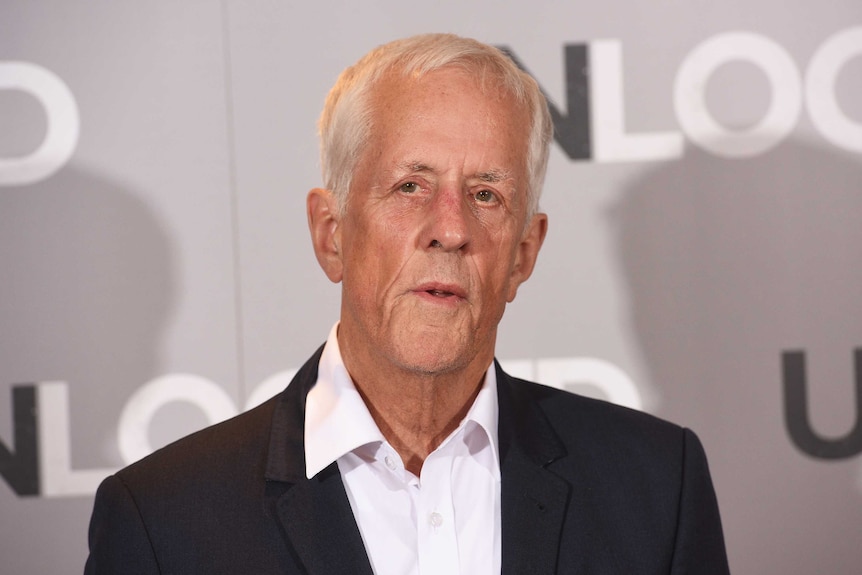 A man in his seventies stands in front of a media wall wearing a black suit jacket and white shirt unbuttoned at the collar.