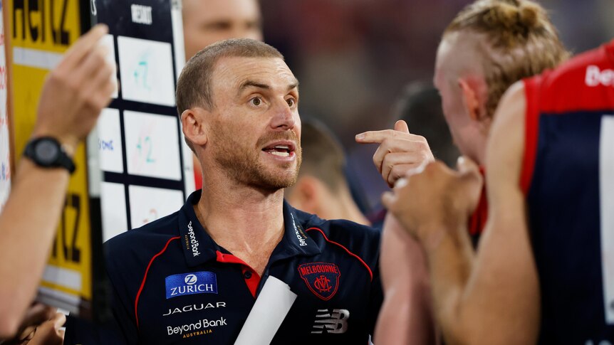 An AFL coach gestures as he stands next to a whiteboard speaking to players during a game. 