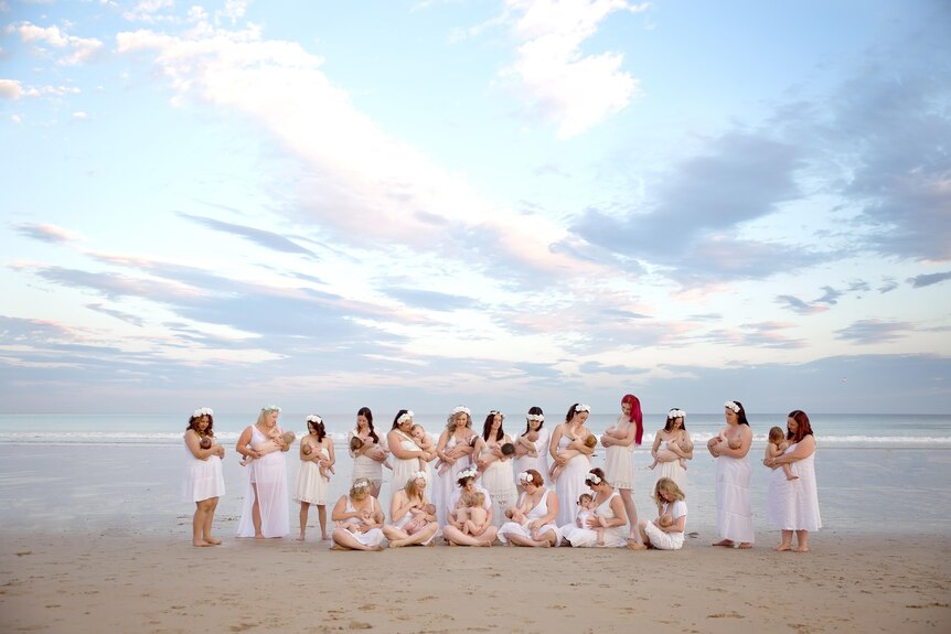 A group of 19 women on a beach breastfeeding their babies
