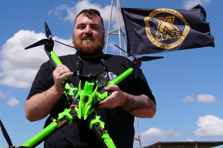 A man in a black t-shirt holds a bright green drone in front of a flag that says 'x-class'