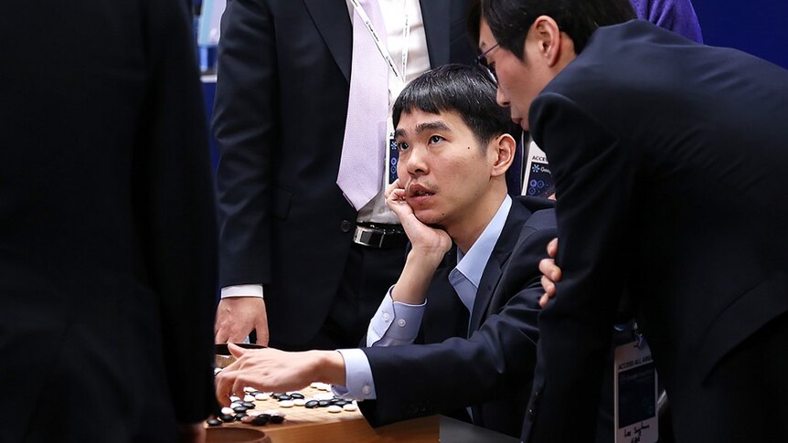 Lee Se-Dol looks up at a man in a suit as he holds his finger on a Go game board