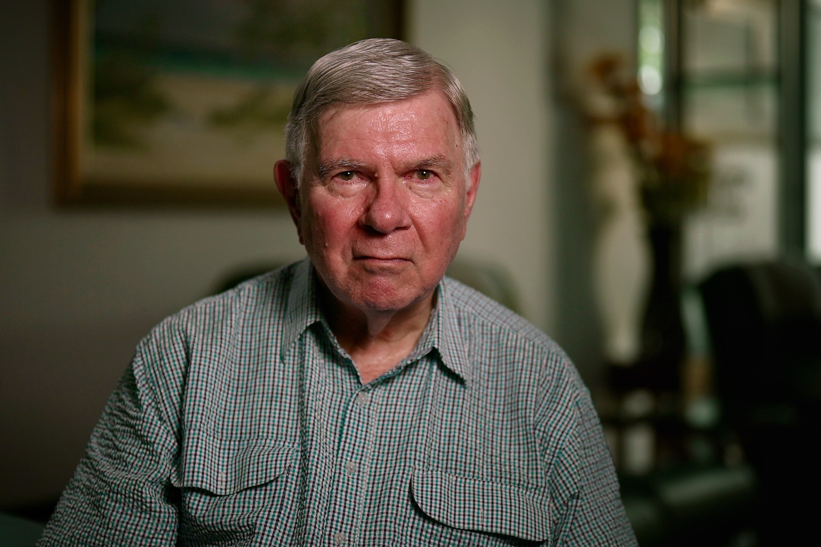 Un hombre mayor, gris, con el pelo corto, vestido con una camisa a rayas y mirando directamente a la cámara, sentado en su casa.