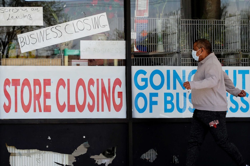 A man in a face mask walks past a small business with a 'store closing' sign on the window
