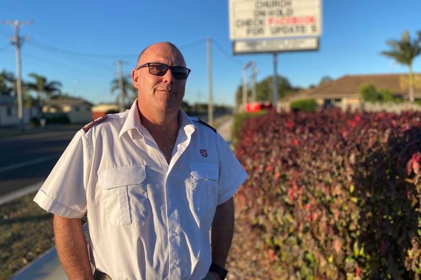 A man in a white shirt stands on a footpath next to a hedge, Salvation Army billboard in the background.