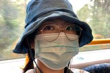 A Chinese woman in a selfie taken on a Melbourne train. She is wearing a floppy hat, large round glasses, and a face mask.