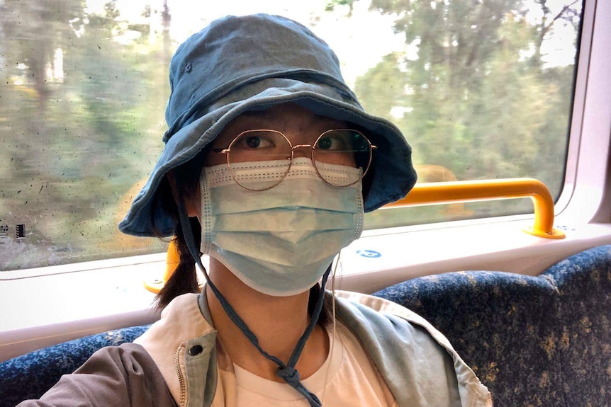 A Chinese woman in a selfie taken on a Melbourne train. She is wearing a floppy hat, large round glasses, and a face mask.