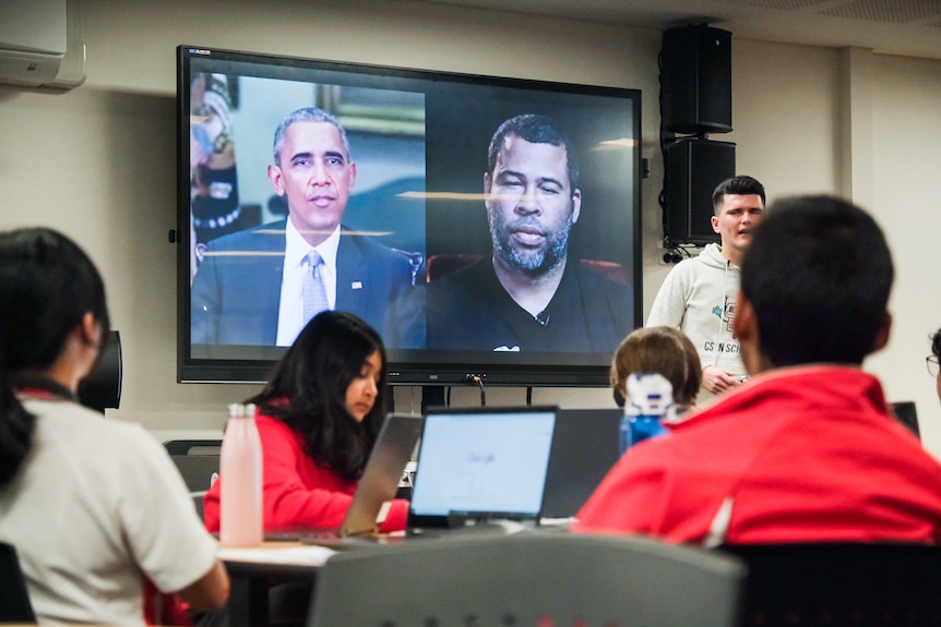 a male teacher standing on front of a classroom with video images behind him