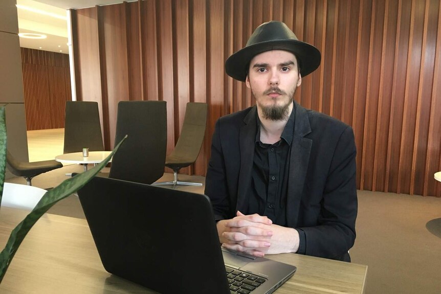 Kayne Weir, wearing a black hat and suit, sits at a desk with a laptop in fornt of him and chairs and a table in the background.