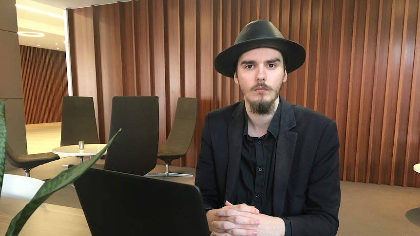 Kayne Weir, wearing a black hat and suit, sits at a desk with a laptop in fornt of him and chairs and a table in the background.