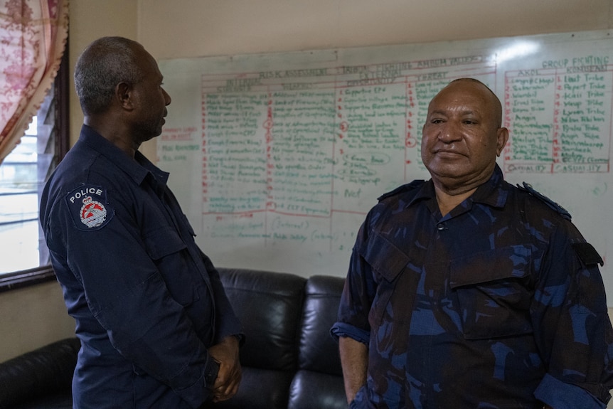 Two police officers standing in front of a whiteboard charting recent tribal fighting.