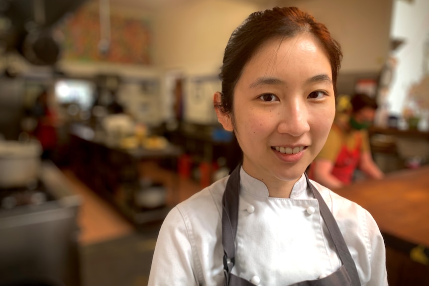 A woman wearing a chef's apron stands in a kitchen.