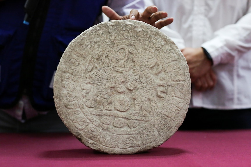 A circular white stone tablet with etchings on it is held up by a human hand as it rests on a pink felt surface.