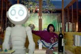 Rashida Jones sits on the couch next to a large white robot.
