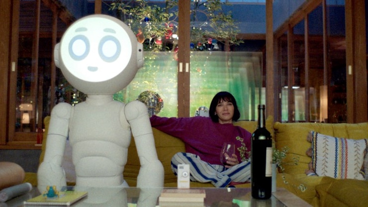 Rashida Jones sits on the couch next to a large white robot.