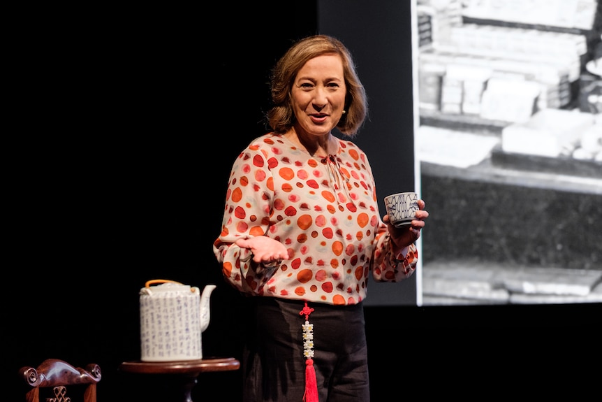 An Asian Australian woman stands on stage, holding a cup of tea in one hand. Behind her is a teapot and a screen.