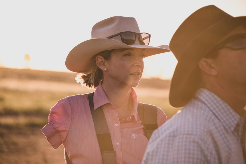 A woman in white brim hat with one arm looks out to distance at sunset 