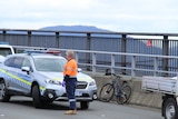 A man wearing high visibility clothing stands near police cars at the scene of a crash involving a bicycle on the Tasman Bridge.