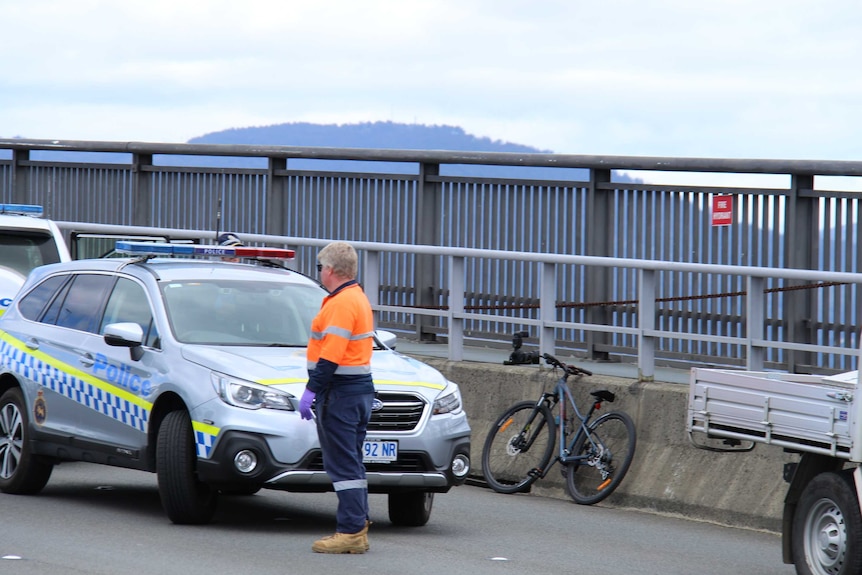 A man wearing high visibility clothing stands near police cars at the scene of a crash involving a bicycle on the Tasman Bridge.