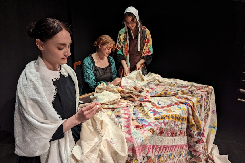 Three female performers in costumes with a quilt.