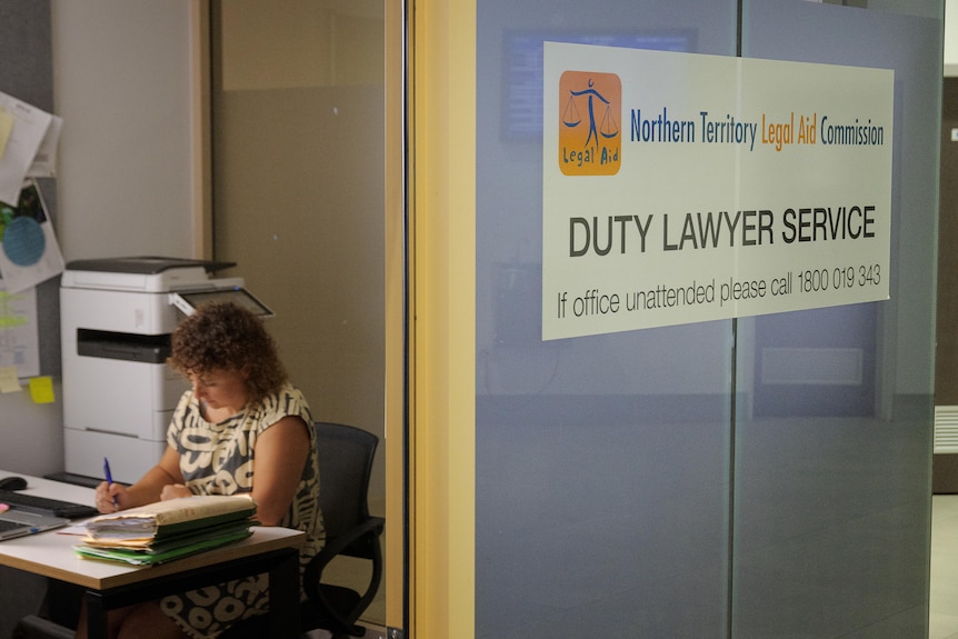 A woman sits at a table and fills in paperwork in an office which reads NT Legal Aid Commission.