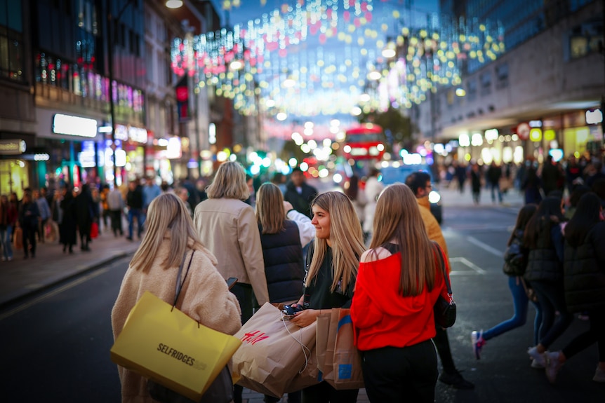 A group of young women with shopping bags walk down a street under a canopy of Christmas lights