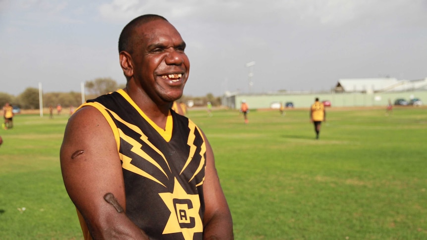 Smiling Indigenous man in a black and yellow football jersey with other players on a field in the background.