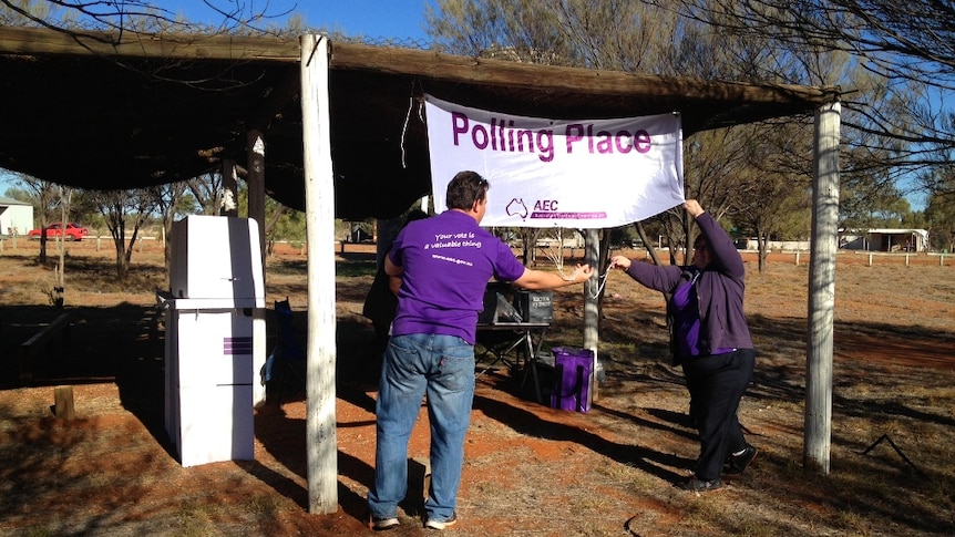 Two people in purple shirts put up a banner which says polling place on it. It's outside and there is red dust.