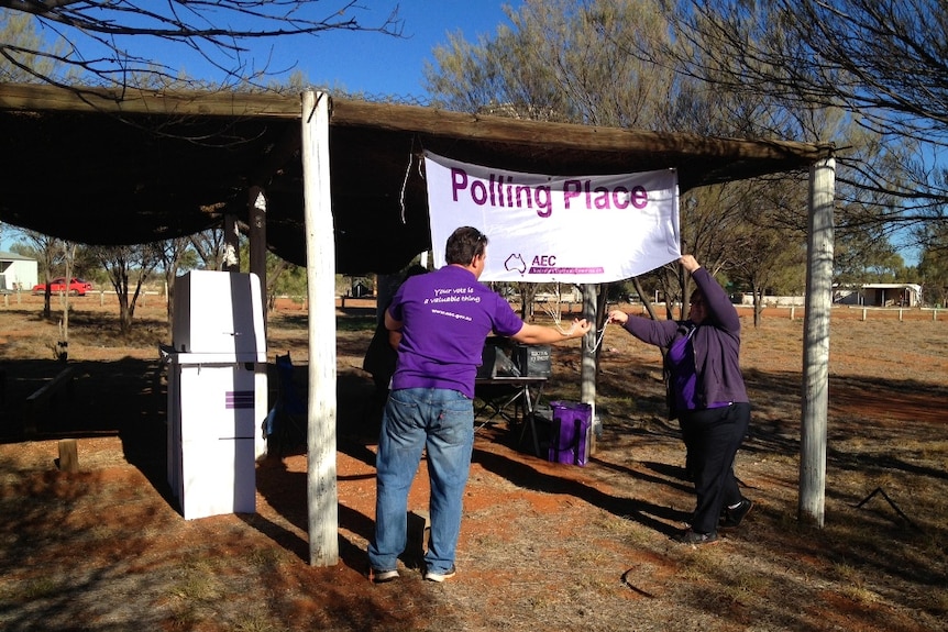 Two people in purple shirts put up a banner which says polling place on it. It's outside and there is red dust.