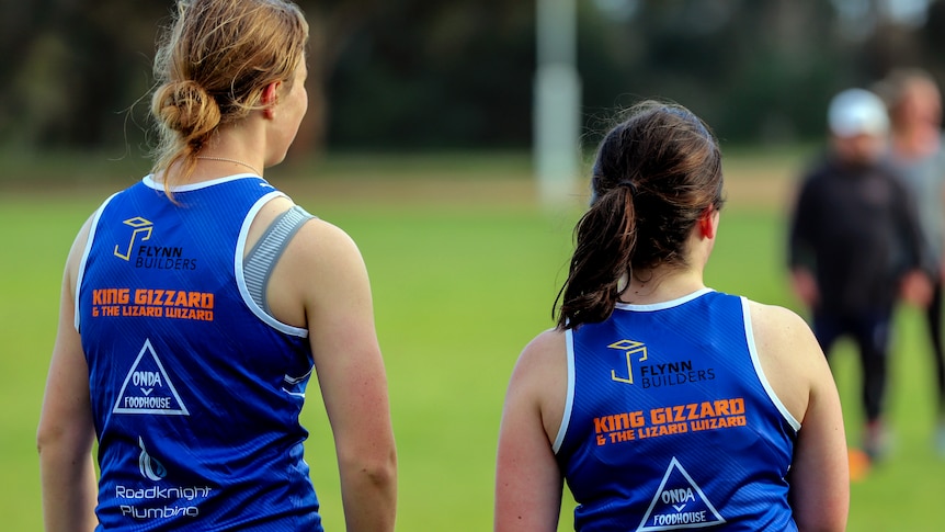 Two women stand on footy oval wearing blue jerseys with graphics on the back