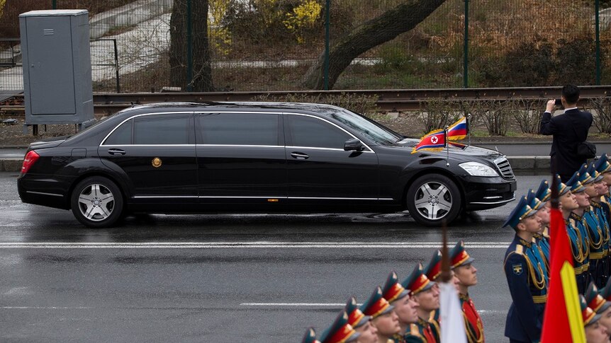 A sleek, stretched black limo bearing North Korean and Russian flags drives past lines of soldiers standing to attention.