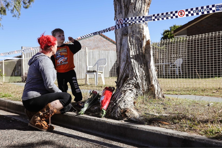 A mother and child lay flowers outside the house, with police tape wrapping around the tree.