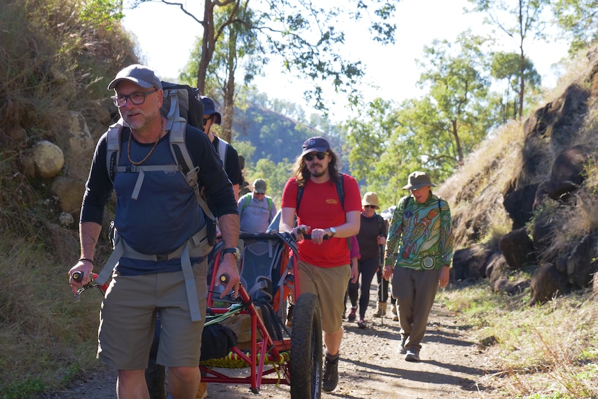 A group of people walk through the countryside, the first two pushing and pulling a cart.