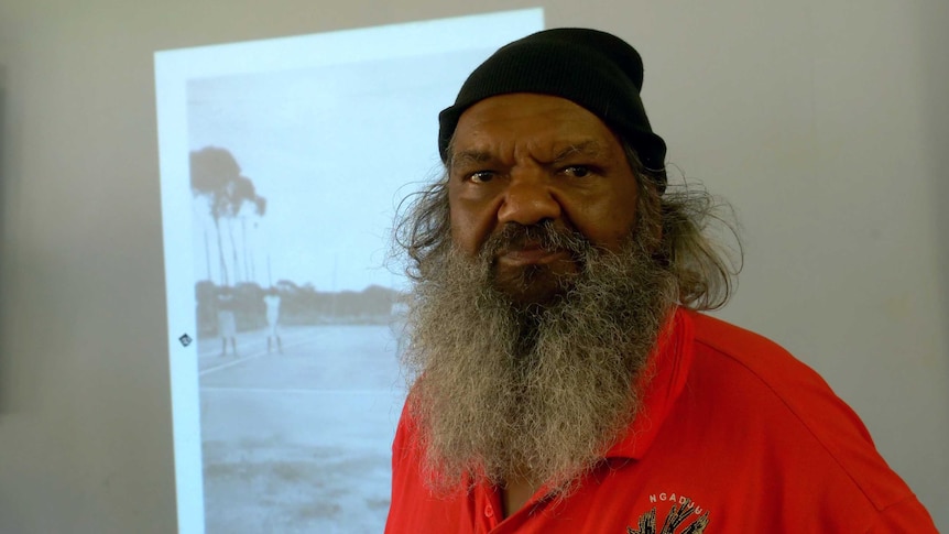 An older Indigenous man with a long beard stands in front of a projected image.