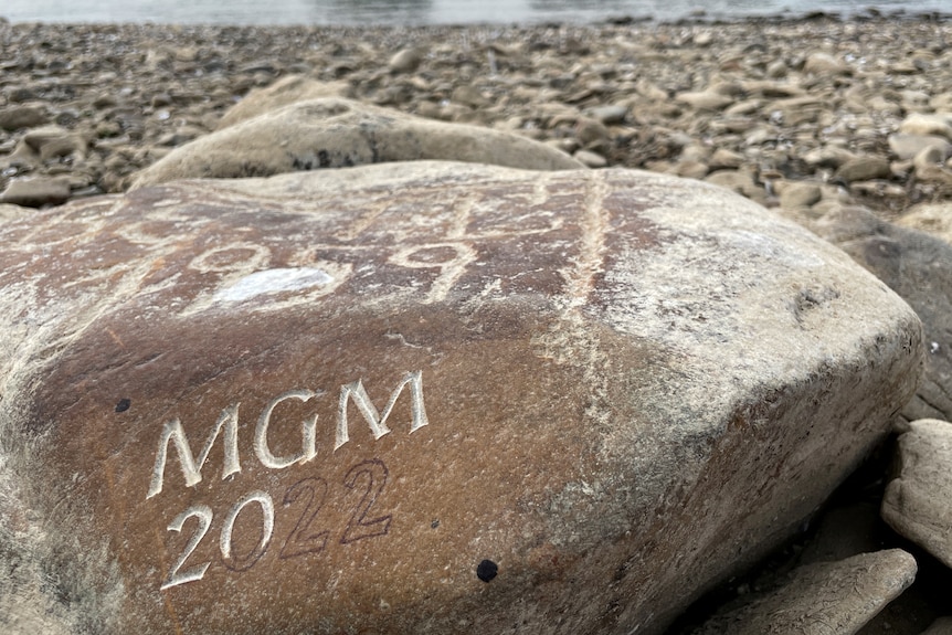  A large rock with an unfinished carving of 'MGM 2022' on it sits on a rocky riverside. River visible in background
