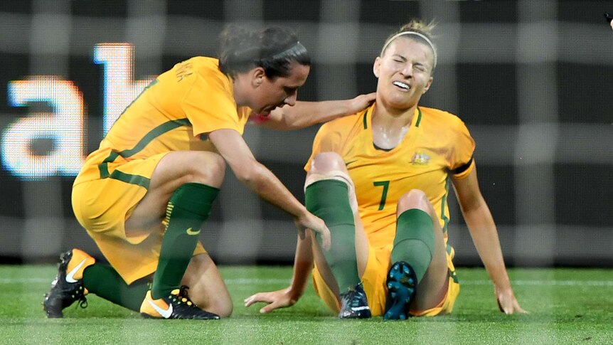 Steph Catley grimaces sitting on the ground as Lisa De Vanna crouches down to check on her