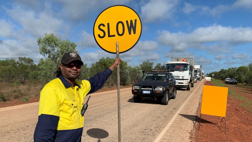 A man holding a sign saying "slow" in front of queuing cars on the highway.