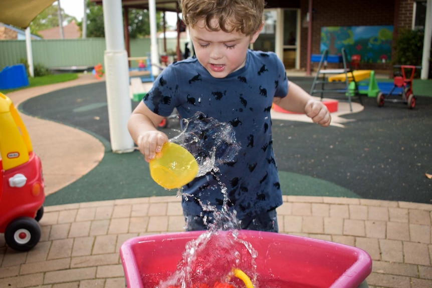 Young boy in blue shirt playing with water at day care centre, colourful toys in background.