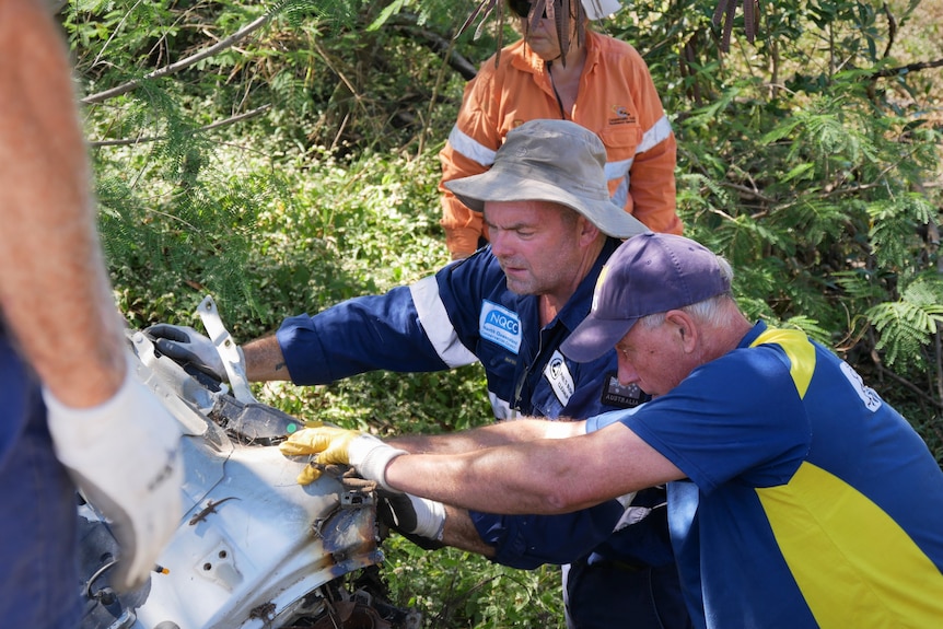 A group of four people wearing gloves and work shirts push a heavy piece of a car out of bush-land.