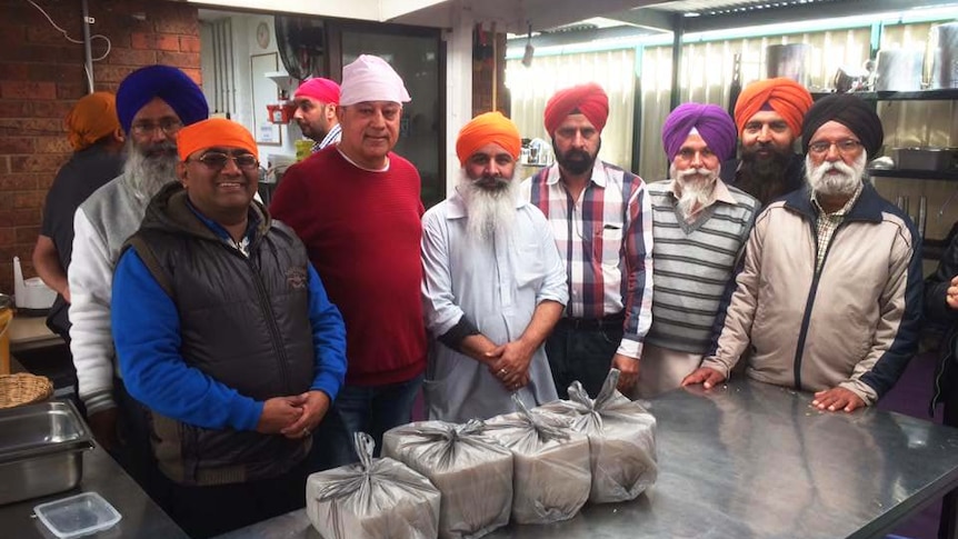 Photo of eight men standing in an industrial kitchen. In front of them is packaged food. 