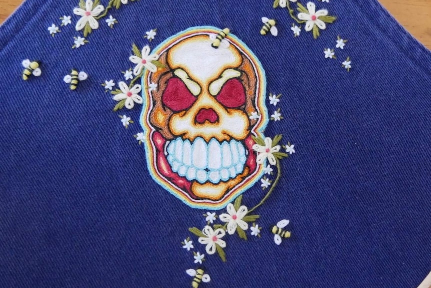 Intricate stitching of embroidered skull on denim, surrounded by embroidered daisies