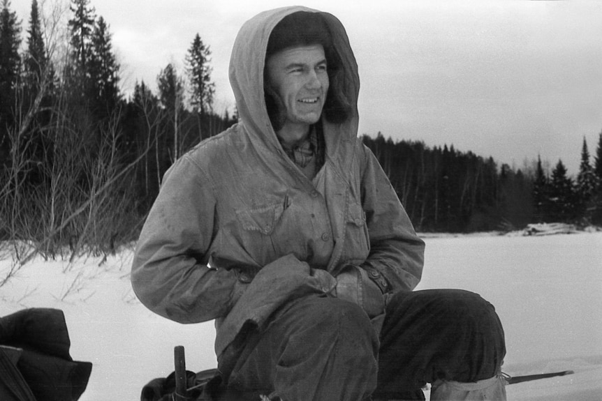 A black and white photo of a smiling young man in a hooded coat sitting in a snowy expanse