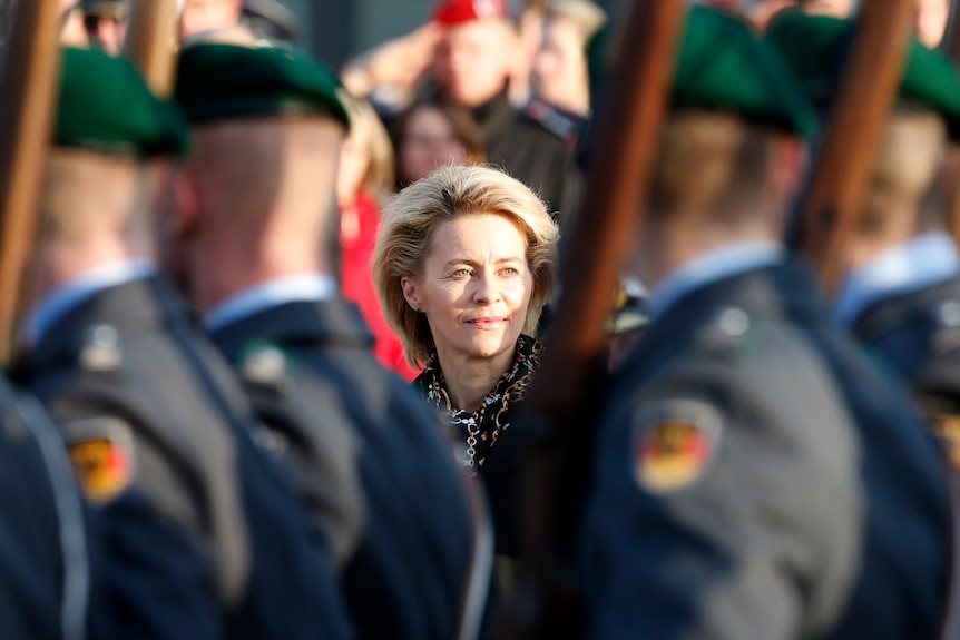 Merkel can be seen in focus in between soldiers who have their backs to the camera who are holding guns over their shoulder